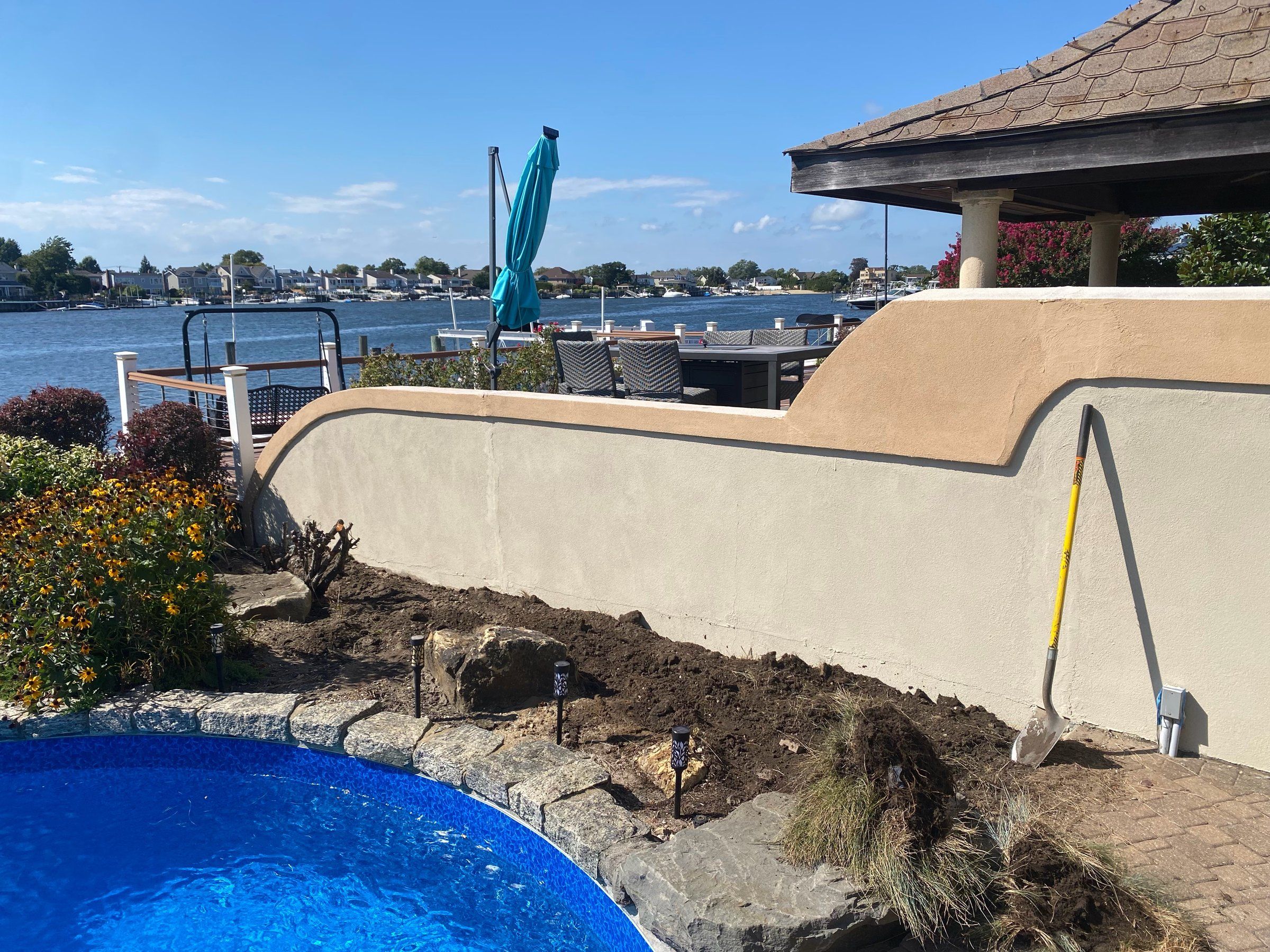 Bare, dug-up planting strip between a pool and a stucco wall on the water