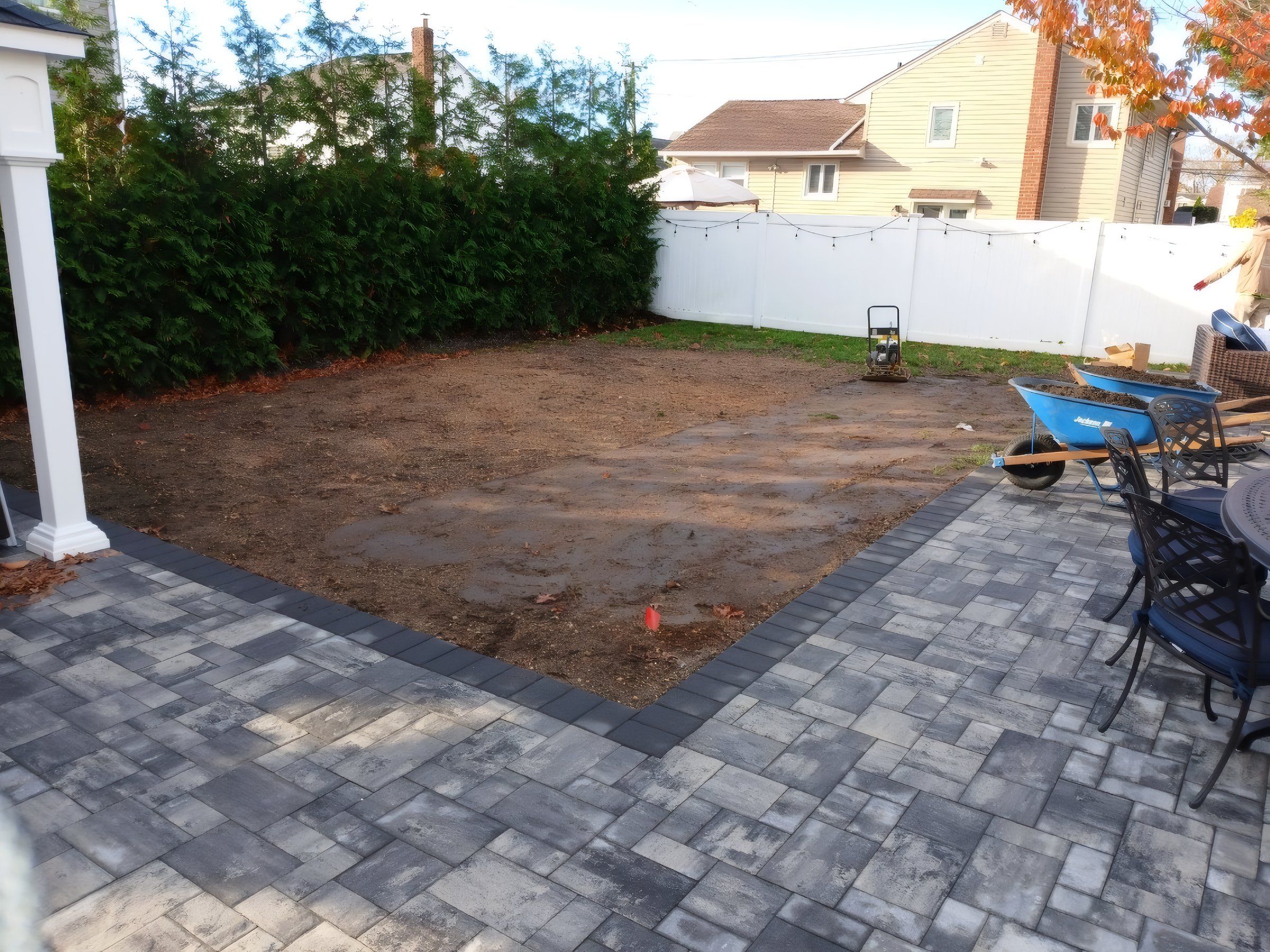Bare soil beside a paver patio and a tall hedge, before sod