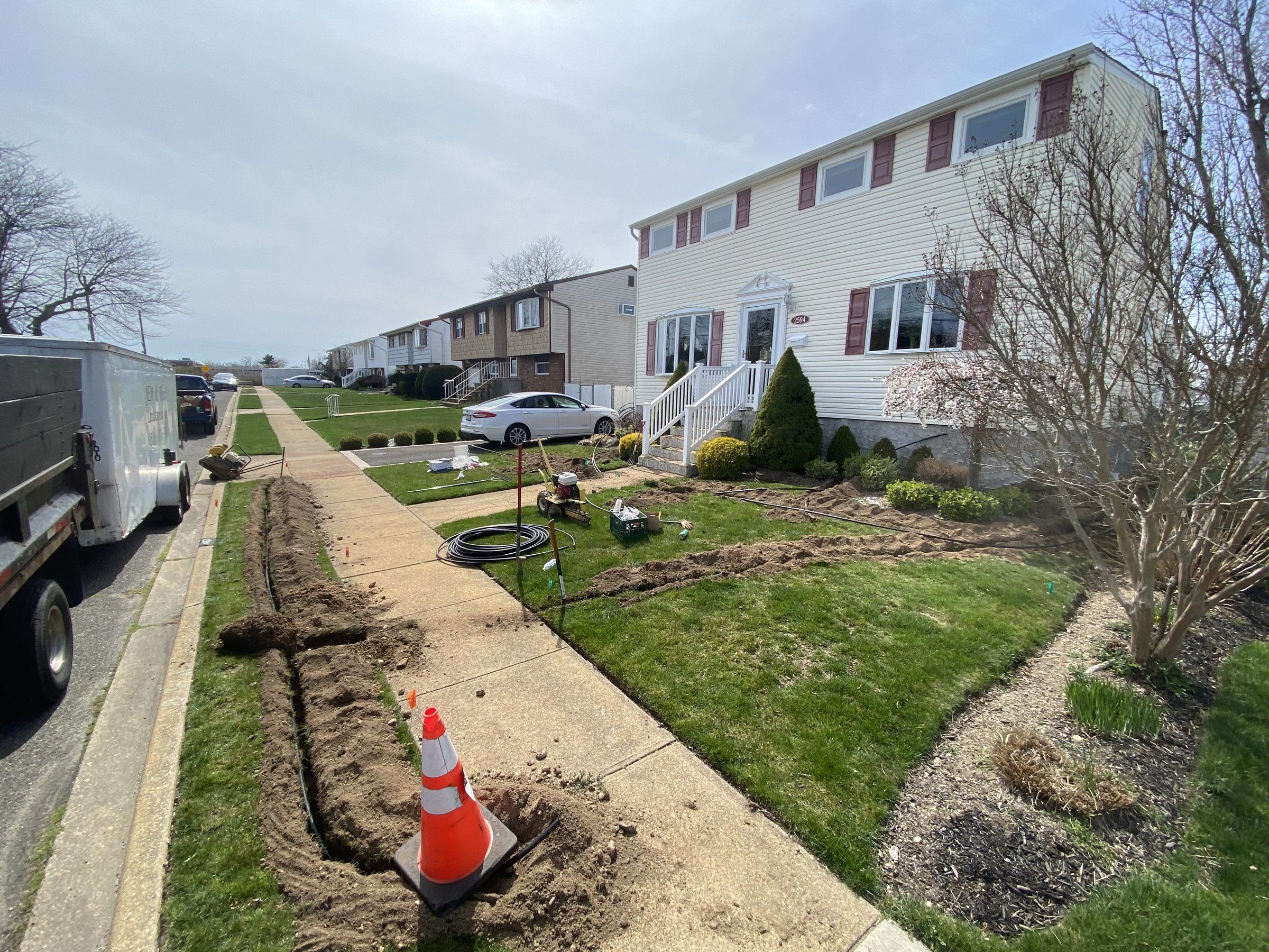 Trench dug along the sidewalk strip, with a coil of pipe on the lawn